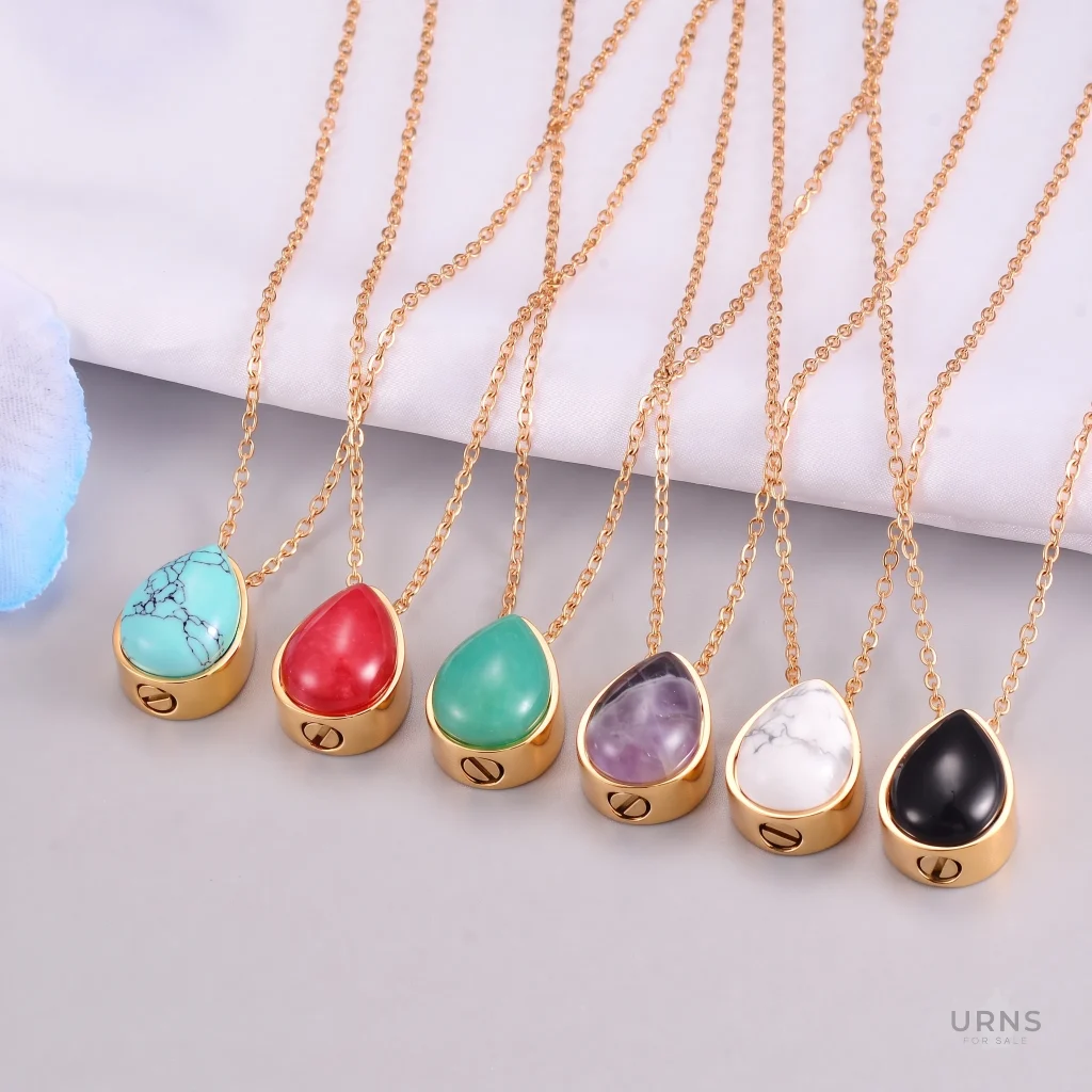 A row of six gold-plated teardrop cremation urn pendants with different gemstone tops (turquoise, red, green jade, amethyst, white marble, black onyx) on chains, shown on a neutral grey and white background with blue detail.