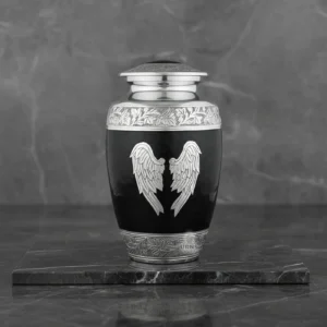 A large black funeral urn for adult ashes, featuring silver angel wings, displayed on a reflective grey marble stand.