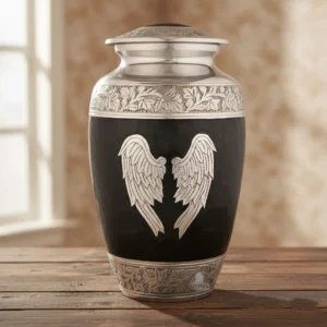 A large black and silver funeral urn for adult human ashes, featuring ornate angel wings, resting on a dark wood surface in a respectful, warm-lit room.
