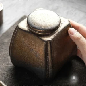 A hand touching the rustic metallic lid of the 'Gilded Earth' ceramic pet urn, showing its tactile, artisan quality and scale.