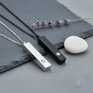 Black and silver Minimalist Bar Urn Necklaces with crystals, placed side-by-side on a dark grey slate background with a white river stone and lavender.