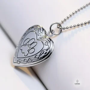 A shiny silver heart-shaped photo locket engraved with a paw print and filigree, on a matching ball chain, for pet memorial.