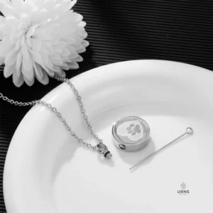 HDR image of the paw print urn necklace kit, showing the pendant, chain, filling funnel, and pin, displayed with a white flower.