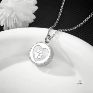 HDR enhanced stainless steel paw print urn necklace for pet ashes, resting on a white plate with a subtle white flower and dark background.