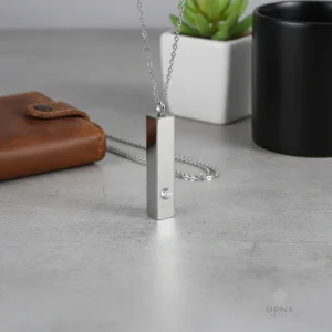 Silver Minimalist Bar Urn Necklace with crystal accent, standing upright on a grey concrete desk next to a leather wallet, showing its suitability for everyday wear.