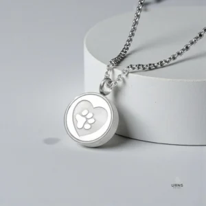 Silver paw print locket necklace for pet ashes, resting on a textured light grey concrete surface in soft, diffused lighting.