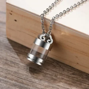 Stainless steel cremation urn necklace with clear vial, shown in a lifestyle setting resting on a dark wooden surface against a light wood box.