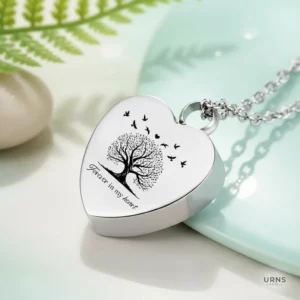 Stainless steel heart cremation urn necklace with Tree of Life and 'Forever in My Heart' engraving, displayed on a tranquil, blue and green background