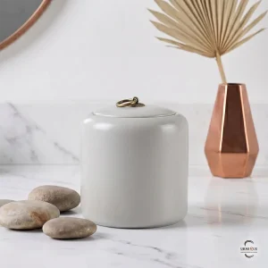 A minimalist memorial display featuring the Alabaster Zen Ceramic Cremation Urn on a white marble surface with smooth river stones and a copper vase.