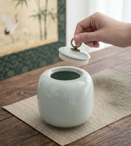 Detailed close-up view of a hand lifting the lid from a light green celadon-style ceramic canister. The lid features an aged bronze ring pull handle and a visible cork liner for an airtight seal. The canister rests on a woven mat on a wooden table, surrounded by tea leaves and pu-erh bricks in an Asian-inspired setting.
