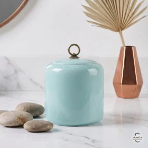A minimalist memorial display featuring the Celeste Zen Ceramic Cremation Urn on a white marble surface with smooth river stones and a copper vase.