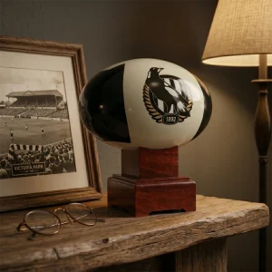 Collingwood Magpies AFL Cremation Urn resting on a rich timber study desk alongside historical Victoria Park memorabilia and a vintage brass lamp.