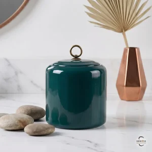 A minimalist memorial display featuring the Emerald Zen Ceramic Cremation Urn on a white marble surface with smooth river stones and a copper vase.
