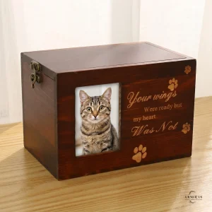 A dark stained wooden keepsake memory box resting on a light wood grain table. The box features an antique brass latch on the side and a portrait of a tabby cat in a photo frame on the front. Engraved in gold to the right of the photo is the poignant text, 'Your wings Were ready but my heart Was Not', accompanied by three engraved paw prints. The background is a softly lit window with a sheer curtain.