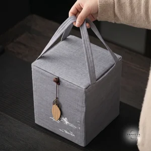 A person's hand holding a closed, square, gray fabric carrying bag with a wooden leaf pendant, resting on a dark wooden table.