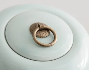 Close-up of a light green ceramic cremation urn lid featuring an antique brass ring pull handle with a decorative sunburst design.
