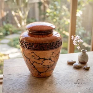 A highly detailed, handcrafted majestic oak wood memorial urn, featuring a two-tone design. The top has a polished domed lid and a dark-stained floral carving band, while the lower body showcases striking, branching Lichtenberg fractal burn patterns. It is presented on a wooden table next to a small vase of cherry blossoms and polished river stones, with a serene garden view through a large window.
