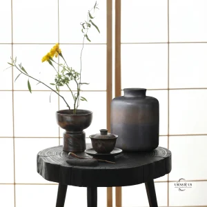 A modern dark textured ceramic urn displayed on a rustic black wooden table alongside a matching dark pottery tea set and yellow wildflowers, set against a traditional softly lit shoji screen
