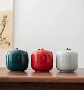 Set of three handcrafted ceramic cremation urns or tea canisters in deep teal green, vibrant red, and light celadon blue, with metal loop-handle lids, on a dark wooden table.