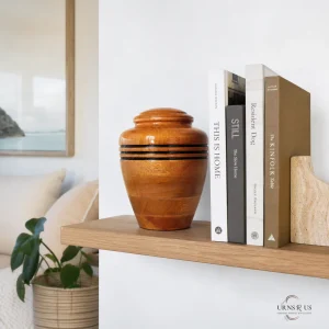 A polished wooden memorial urn with three black bands sitting on a floating wooden shelf next to a stack of books in a contemporary living room setting.