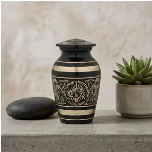 A small, black and gold brass cremation keepsake urn with an ornate etched design sits on a concrete shelf next to a smooth dark stone and a potted succulent.