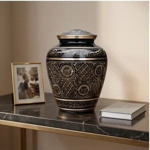 A black and gold brass cremation urn sits on a marble table next to a gold-framed photo of a smiling person and a closed book.