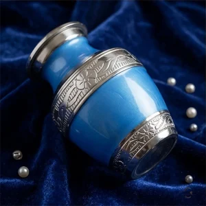 A blue sapphire cremation urn with intricate silver skirting, shown on a dark blue velvet cloth with scattered pearls.