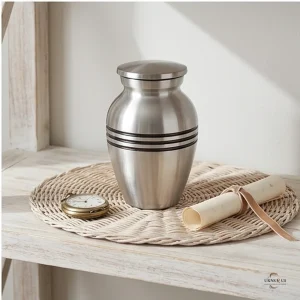 A classic silver brass keepsake urn for ashes is displayed on a woven mat alongside a vintage pocket watch and a rolled scroll tied with ribbon, set against a neutral background.