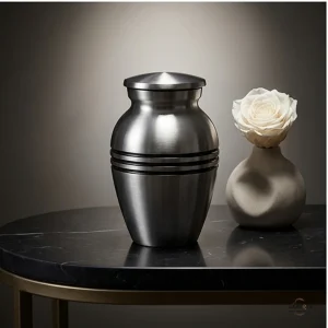 A classic silver brass keepsake urn for ashes sits on a dark marble table next to a vase with a white rose.