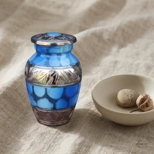 A small, ornate blue keepsake urn with silver accents sits on a textured linen tablecloth next to a small ceramic bowl containing two smooth stones and a dried seed pod. The urn has a lid with a blue jewel and intricate silver detailing around its base and neck.