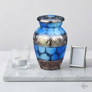 A blue and silver keepsake urn for ashes, labeled 'Cloud of Peace', sits on a marble slab next to a lit tea candle in a glass holder and a small silver photo frame.