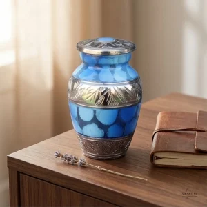 A blue and silver keepsake urn with a circular pattern sits on a wooden table next to a small leather-bound journal and a sprig of lavender.