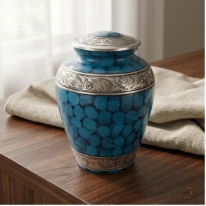 A close-up of the Cynosure Sea Blue Cremation Urn, featuring a textured blue surface with darker oval shapes and ornate silver bands, placed on a wooden table next to a beige cloth.
