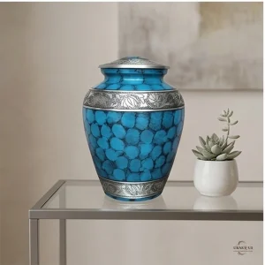 A blue cremation urn with a patterned silver band sits on a glass shelf next to a small succulent plant.