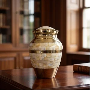 A brass cremation urn with a mother-of-pearl inlay and gold bands, sitting on a polished wooden desk in a library setting.