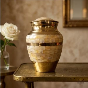 A Pearl Petal Adult Brass Cremation Urn for Ashes sits on a brass table next to a white rose and a glass vase.