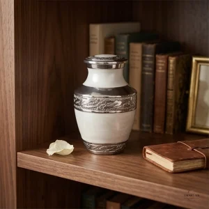 A pearl white and silver keepsake cremation urn with intricate detailing sits on a wooden shelf next to a small stack of books and a leather-bound journal.