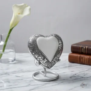 A silver heart-shaped keepsake urn with intricate floral engraving, displayed on a marble desk next to a white calla lily and two leather-bound books. The urn is mounted on a silver stand.