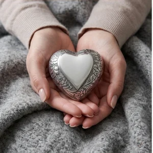 A pair of hands gently holding a silver heart-shaped keepsake urn adorned with intricate floral engravings, resting on a textured grey fabric.