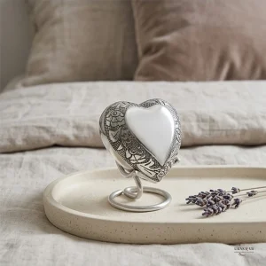 A Satori silver heart-shaped keepsake urn for ashes, displayed on a round tray with lavender sprigs, placed on a bed with soft linen bedding and pillows.