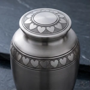 Close-up view of a silver cremation urn with a decorative band of hearts around the body and lid.