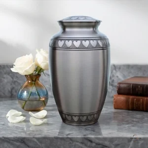 A silver cremation urn decorated with a band of hearts sits on a marble surface next to a vase of white roses and two old books.