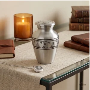A silver keepsake cremation urn with a heart pattern sits on a table runner next to a lit candle, a locket, a leather-bound journal, and a stack of old books.