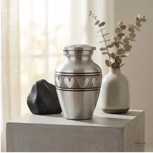 A silver cremation urn with a heart engraving, placed next to a dark stone and a vase of eucalyptus on a concrete pedestal.