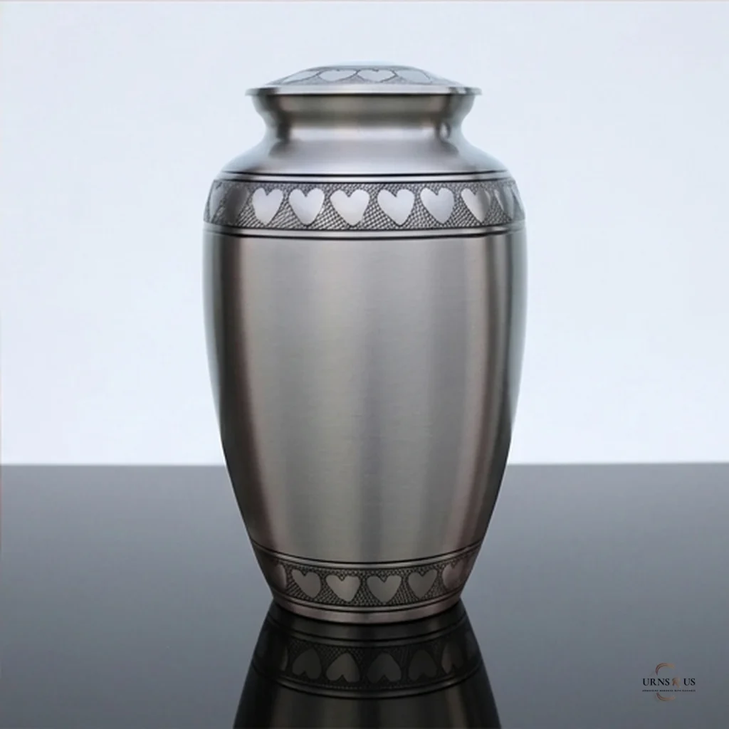 A silver cremation urn with a heart pattern on the lid and around the top and bottom edges, placed on a reflective surface against a white background.