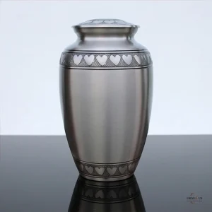 A silver cremation urn with a heart pattern on the lid and around the top and bottom edges, placed on a reflective surface against a white background.