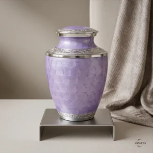 A lavender cremation urn with a textured pattern and silver trim, displayed on a silver stand in front of a draped fabric.