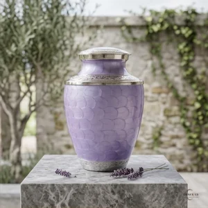 A detailed mockup of the Silver Lining Lavender Adult Large Cremation Urn for Ashes, featuring a beautiful lavender-colored urn with intricate silver detailing, placed on a marble surface with lavender sprigs in front of a stone wall and olive tree.
