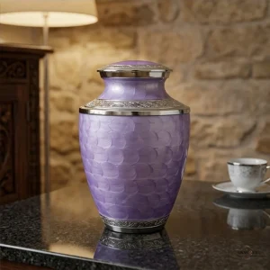 A large, ornate lavender cremation urn with intricate silver trim and a patterned surface, displayed on a dark, reflective table next to a delicate teacup and saucer. The background features a rustic stone wall and a wooden cabinet.