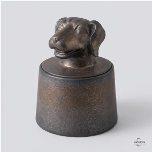 A close-up front view of a handcrafted stoneware cremation urn for ashes, shaped like a dog's head on a cylindrical base. The urn has a bronze and dark grey speckled finish.